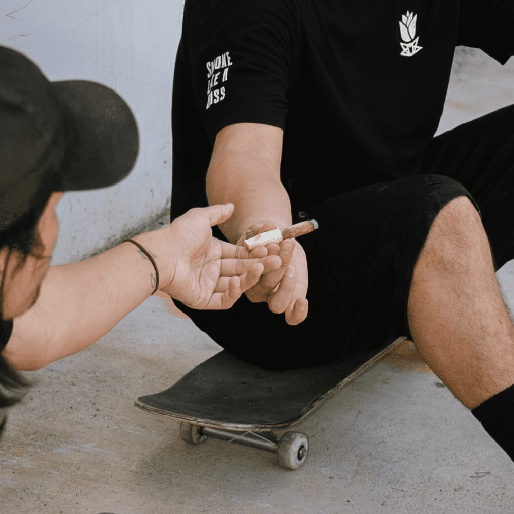 Someone on a skateboard passes a long burning cannabis cigar to an outstretched hand.