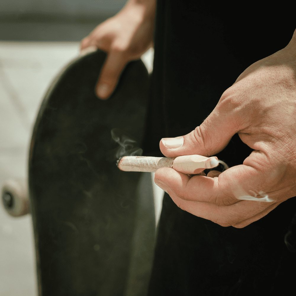 A close up of a person’s hands holding a skateboard in the background and smoking a blunt wrapped in hemp paper and completed with a wood blunt tip