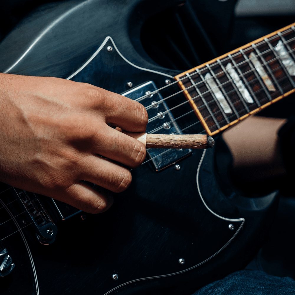 A close up of hands holding a black electric guitar and a slow-burning cannabis cigar.