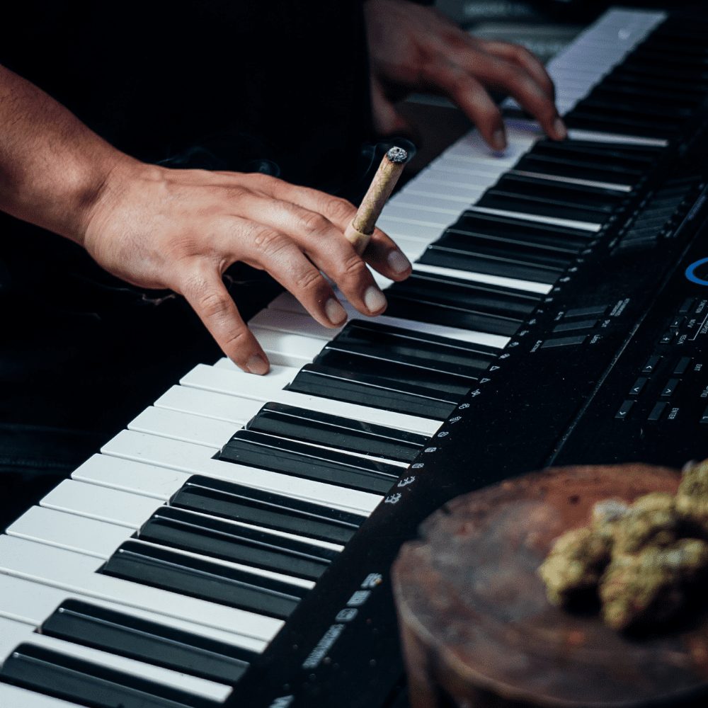 a close up of hands holding a personal-sized 2-4g hemp-leaf wrapped cannagar while they play the piano. Weed is displayed in the foreground.