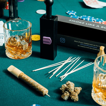 Cannagar hemp blunt laying on a felted gambling table next to weed nugs, bamboo skewers, cannagar box set, and whiskey.