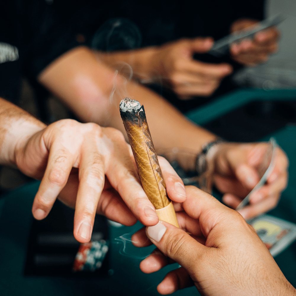 Close up of hands passing a large, hemp leaf wrapped blunt as they play a card game at a table with poker chips. The blunt has a wooden filter tip.