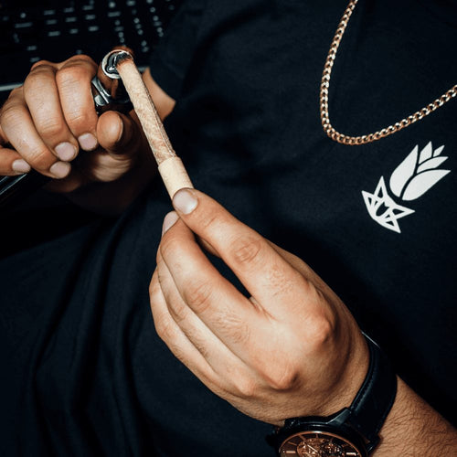 A close up of hands lighting a cannagar, a hemp-wrapped blunt, that’s larger than a preroll. The model wears a black shirt with a white Purple Rose Supply logo.